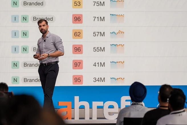 A man in a checkered shirt presents data on a large screen, showing "Branded" keywords, numbers (53, 96, 62, 97, 95), millions (75M, 71M, 57M, 55M, 41M, 34M), and line graphs. The Ahrefs logo is below the screen.