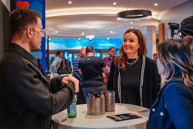 Three people are conversing around a high-top table in a bustling indoor setting. A man in glasses is on the left, looking at two women on the right. One woman smiles while another looks at her.